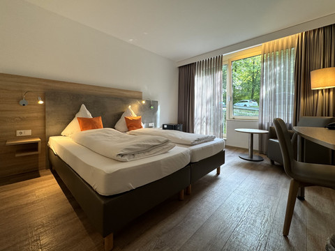 Innenansicht Doppelzimmer - Waldhotel Tropfsteinhöhle Helles Hotelzimmer mit zwei Einzelbetten, Holzfußboden, Schreibtisch und Blick auf ein Waldstück.