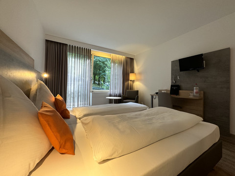 Innenansicht Doppelzimmer - Waldhotel Tropfsteinhöhle Doppelzimmer mit zwei weißen Betten, hellen Wänden, Flachbildfernseher, Fenster mit Blick ins Grüne.