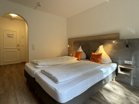 Innenansicht Doppelzimmer - Waldhotel Tropfsteinhöhle Helles Doppelzimmer mit zwei weißen Betten, Holzboden, Akzentkissen und Türbogen im Hintergrund.