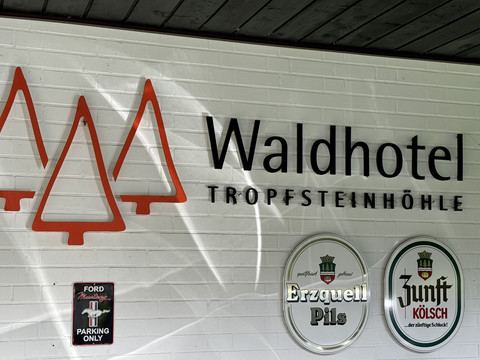 Außenansicht Waldhotel Tropfsteinhöhle Logo des Waldhotel Tropfsteinhöhle an einer weißen Wand, umgeben von zwei Biermarken-Schildern.