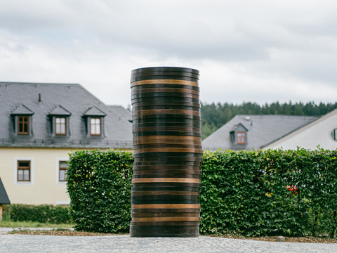 PurplePath_ Coin Stack 2_Sean Scully in Schneeberg_Kunstwerk 2_Foto_ Johannes Richter.jpg Eine massive, bronzefarbene Skulptur vor grüner Hecke und Gebäuden im Hintergrund.A massive, bronze-colored sculpture in front of a green hedge and buildings in the background.Masivní bronzová socha před zeleným živým plotem a budovami v pozadí.Masywna rzeźba w kolorze brązu na tle zielonego żywopłotu i budynków w tle.Een massief, bronskleurig beeld voor een groene haag en gebouwen op de achtergrond.Una massiccia scultura color bronzo davanti a una siepe verde e a edifici sullo sfondo.
