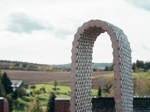 PurplePath_ Bogen_Leunora Salihu in der Gedenkstätte Hoheneck _ Stollberg _Werk_Foto_ Ernesto Uhlmann(1).jpg Ein Skulpturenbogen aus gleichmäßigen, kreisförmigen Elementen, vor einer grünen Landschaft.A sculptural arch of regular, circular elements in front of a green landscape.Sochařský oblouk z pravidelných kruhových prvků před zelenou krajinou.Rzeźbiarski łuk z regularnych, okrągłych elementów na tle zielonego krajobrazu.Een sculpturale boog van regelmatige, cirkelvormige elementen voor een groen landschap.Un arco scultoreo di elementi regolari e circolari davanti a un paesaggio verde.