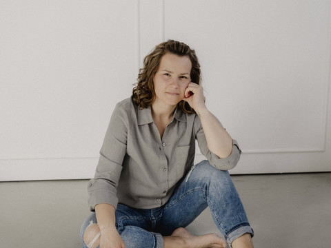 Annette Müller Eine Frau in Jeans und grauer Bluse sitzt barfuß entspannt auf dem Boden in hellem Raum.