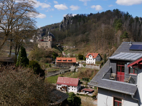 Blick auf eine mittelalterliche Burg in hügeliger Landschaft mit Fachwerkhäusern und Bäumen, unter blauem Himmel mit wenigen Wolken.