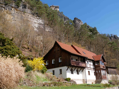 Ein Fachwerkhaus mit rotem Dach steht vor einer felsigen Klippe, umgeben von Bäumen und gelbem Strauch, unter klarem, blauem Himmel.