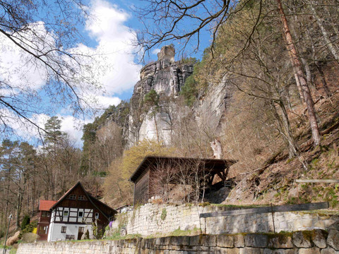 Fachwerkhaus mit rotem Dach vor steilem Felsen und bewaldetem Hang, unter blauem Himmel mit weißen Wolken.