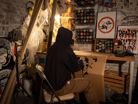 Banksy bei der Arbeit - House of Banksy Leipzig In der Ausstellung des House of Banksy in Leipzig steht eine Werkbank mit Spraydosen, an der eine vermummte Puppe sitzt, die den unbekannten Künstler Banksy bei der Arbeit darstellen sollIn the exhibition at the House of Banksy in Leipzig, there is a workbench with spray cans at which sits a masked doll that is supposed to represent the unknown artist Banksy at workNa výstavě v Domě Banksyho v Lipsku je pracovní stůl s plechovkami od sprejů, u kterého sedí maskovaná loutka, která má představovat neznámého umělce Banksyho při práci.Na wystawie w Domu Banksy'ego w Lipsku znajduje się stół warsztatowy z puszkami ze sprayem, przy którym siedzi zamaskowana lalka, która ma reprezentować nieznanego artystę Banksy'ego przy pracyIn de tentoonstelling in het Huis van Banksy in Leipzig staat een werkbank met spuitbussen waarop een gemaskerde pop zit die de onbekende kunstenaar Banksy aan het werk moet voorstellen.Nella mostra della Casa di Banksy a Lipsia c'è un banco di lavoro con bombolette spray su cui siede un pupazzo mascherato che dovrebbe rappresentare l'artista sconosciuto Banksy al lavoro.