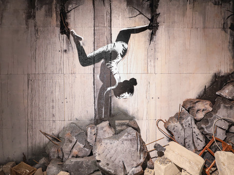 Obrázek z výstavy v Banksyho domě v Lipsku, na kterém je graffiti gymnastky ve stojce, které umělec nastříkal na trosky na Ukrajině.