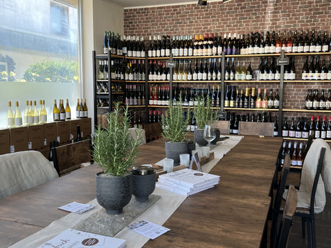 Weinhandel Kork & Kante Modernes Weinlokal mit hölzernem Tisch, Kräutertöpfen und Weinflaschenregalen an der Backsteinwand.