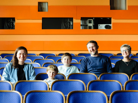 Technische Sammlungen der Stadt Dresden Eine Familie sitzt lächelnd in einem leeren Kinosaal mit orangen Wänden und blauen Sitzen.A family sits smiling in an empty movie theater with orange walls and blue seats.V prázdném kině s oranžovými stěnami a modrými sedadly sedí usměvavá rodina.Rodzina siedzi uśmiechnięta w pustym kinie z pomarańczowymi ścianami i niebieskimi fotelami.Een familie zit glimlachend in een lege bioscoop met oranje muren en blauwe stoelen.Una famiglia siede sorridente in un cinema vuoto con pareti arancioni e poltrone blu.