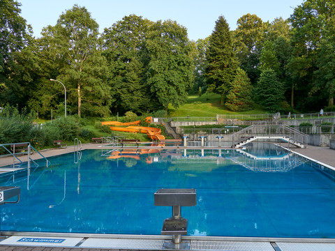 Ein leeres Schwimmbecken mit blauer Wasseroberfläche, umgeben von grünen Bäumen und einer orangefarbenen Rutsche im Hintergrund.