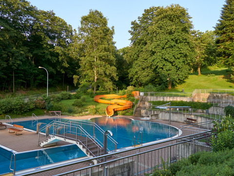 Stadtbad Hohnstein Ein Freibad mit blauem Wasser, einer gelben Rutsche und umgeben von grünen Bäumen und Rasenflächen bei sonnigem Wetter.