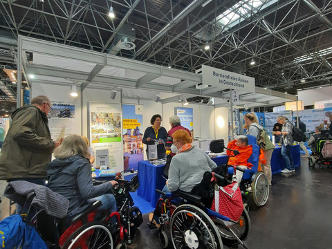 REHACARE Düsseldorf2024_Messestand.JPG Messestand mit mehreren Personen in Rollstühlen, die sich Informationsmaterial ansehen.