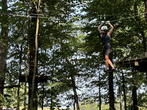 Kletterwald Königstein: Person in schwarzer Kleidung und Helm balanciert auf einem Seil im Kletterpark, umgeben von grünen Bäumen bei sonnigem Wetter.
