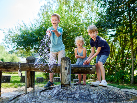 Drei Kinder spielen fröhlich an einem Wasserspielplatz, spritzen Wasser und lachen, umgeben von grünen Bäumen und sonnigem Wetter.Three children play happily at a water playground, splashing water and laughing, surrounded by green trees and sunny weather.Tři děti si spokojeně hrají na vodním hřišti, cákají vodu a smějí se, obklopené zelenými stromy a slunečným počasím.Trójka dzieci bawi się radośnie na wodnym placu zabaw, chlapiąc wodą i śmiejąc się, otoczona zielonymi drzewami i słoneczną pogodą.Drie kinderen spelen vrolijk op een waterspeelplaats, spetteren water en lachen, omringd door groene bomen en zonnig weer.Tre bambini giocano felici in un parco giochi acquatico, spruzzando acqua e ridendo, circondati da alberi verdi e da un tempo soleggiato.