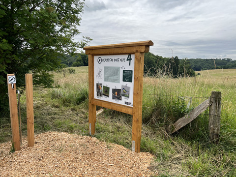 Hexenpfad Dorf Olpe Informationstafel einer Wanderroute in ländlicher Landschaft mit Feldern und bewölktem Himmel.