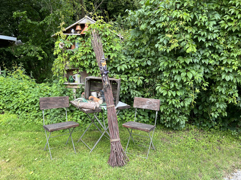 Hexenpfad Dorf Olpe Garten mit zwei Holzstühlen, einem Tisch, dekorativen Pflanzentöpfen und grüner Blatthecke im Hintergrund.