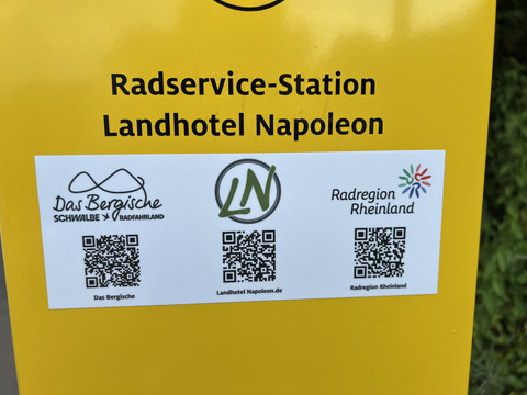 Radservice-Station Gelbes Schild mit Aufschrift "Radservice-Station Landhotel Napoleon" und drei QR-Codes darunter.