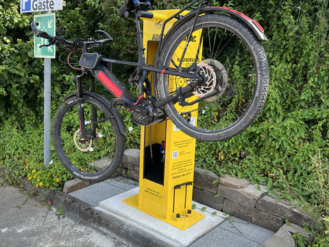 Radservice-Station Fahrrad steht aufrecht in einer gelben Wartungsstation vor grüner Vegetation und einem Parkplatzschild.