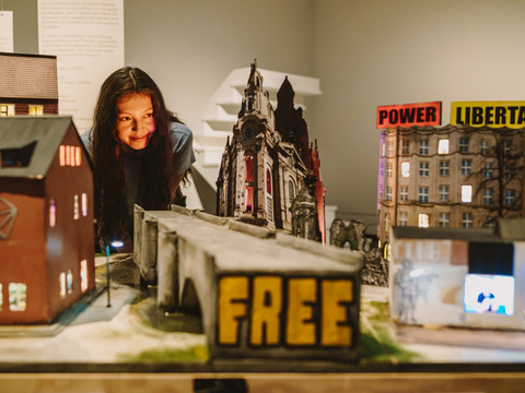 Freiheit 9_Isabel Noack.jpg Ein Mädchen betrachtet ein Diorama mit Miniaturgebäuden und dem Wort "FREE".A girl looks at a diorama with miniature buildings and the word "FREE".Dívka se dívá na dioráma s miniaturními budovami a nápisem "FREE".Dziewczynka patrzy na dioramę z miniaturowymi budynkami i napisem "FREE".Een meisje kijkt naar een kijkdoos met miniatuurgebouwen en het woord "VRIJ".Una ragazza guarda un diorama con edifici in miniatura e la scritta "FREE".