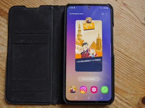 Smartphone in schwarzer Hülle auf Holztisch zeigt eine App mit dem Titel "Hörerlebnispfad Pirna" und einer Illustration von zwei Personen vor Gebäuden.