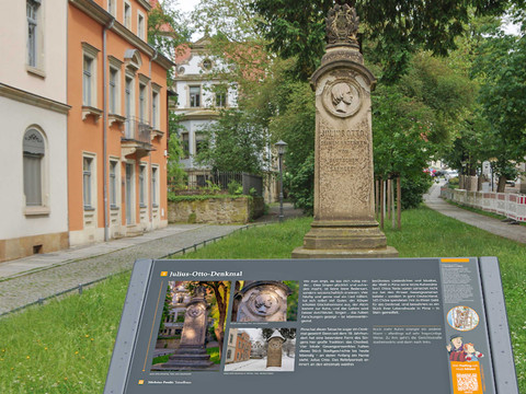 Steinernes Julius-Otto-Denkmal mit Relief in einem grünen Park, daneben ein Informationsschild. Im Hintergrund sind historische Gebäude zu sehen.