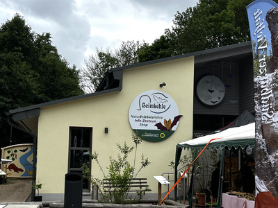 Gelbes Gebäude des Natur- und Erlebniszentrums Heimkehle mit Schild, Bänken und umgeben von Grün.