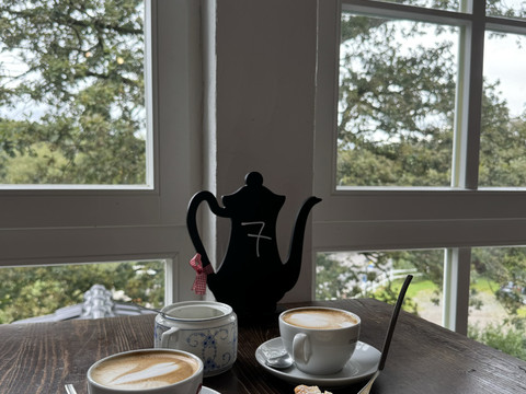 Kaffee & Käsekuchen Kaffeeszene mit Cappuccinos, Teekanne und Gebäck, vor großen Fenstern mit grünem Ausblick.