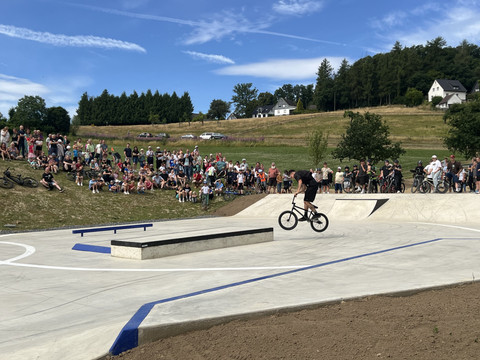 Skate und Bikepark Waldbröl Eine große Menschengruppe beobachtet BMX-Fahrer auf einer Beton-Skateanlage vor grüner Landschaft.