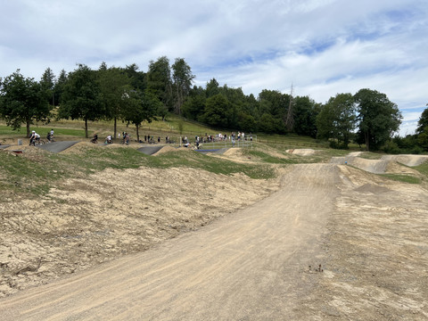 Skate und Bikepark Waldbröl Mehrere Fahrradfahrer auf einer hügeligen BMX-Strecke inmitten bewaldeter Landschaft und blauem Himmel.