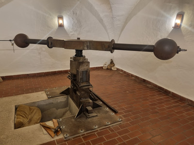 Ein massives, historisches Hebewerk aus Metall steht in einem Gewölbekeller mit Backsteinboden.