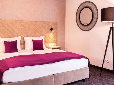 Victor’s Residenz-Hotel Gummersbach Modern eingerichtetes Hotelzimmer mit Doppelbett, beigen Kopfteil, lila Akzenten und Stehleuchte.