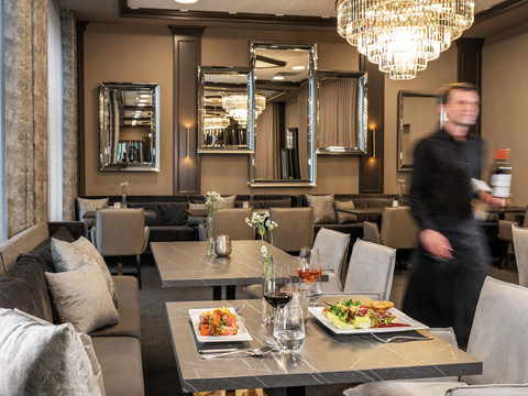 Victor's Residenz-Hotel Gummersbach Ein stilvoll eingerichtetes Restaurant mit gedeckten Tischen, elegantem Kronleuchter und vorbeigehendem Kellner.