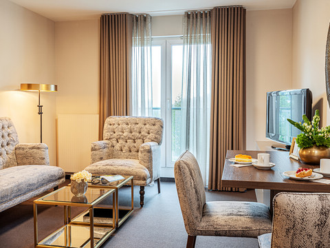 Victor's Residenz-Hotel Gummersbach Gemütliches Wohnzimmer mit zwei Polstersesseln, Couchtisch, Esstisch mit Sesseln und TV an der Wand.