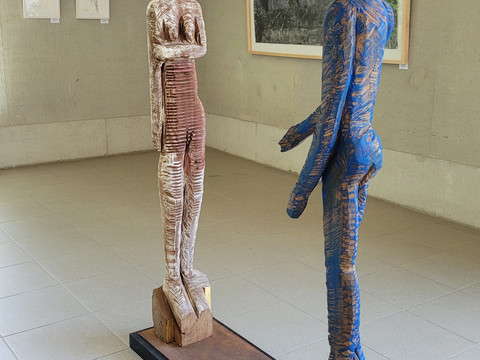 Begegnung©GeraldWunderlich.jpg Zwei farbige Holzfiguren stehen sich in einer Galerie gegenüber, umrahmt von Kunstwerken an den Wänden.
