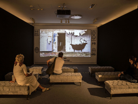 Drei Personen betrachten eine Projektion eines Graffitis im Banksy-Stil in einem dunklen Raum.Three people look at a projection of Banksy-style graffiti in a dark room.Tři lidé se dívají na projekci graffiti ve stylu Banksyho v temné místnosti.Trzy osoby patrzą na projekcję graffiti w stylu Banksy'ego w ciemnym pokoju.Drie mensen kijken naar een projectie van Banksy-achtige graffiti in een donkere kamer.Tre persone guardano una proiezione di graffiti in stile Banksy in una stanza buia.