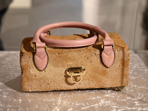 Eine Handtasche mit rosa Griffen auf einem Ziegelstein, präsentiert in einer Ausstellung.A handbag with pink handles on a brick, presented in an exhibition.Kabelka s růžovými uchy na cihle, prezentovaná na výstavě.Torebka z różowymi uchwytami na cegle, prezentowana na wystawie.Een handtas met roze hengsels op een baksteen, gepresenteerd in een tentoonstelling.Una borsa con manici rosa su un mattone, presentata in una mostra.