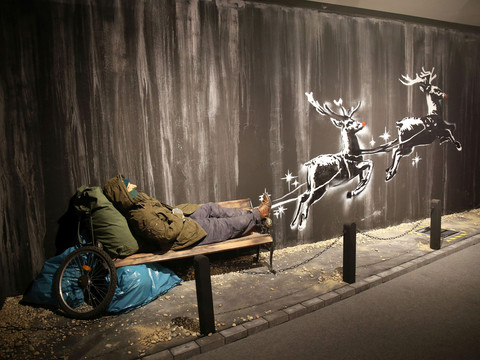 12 House of Banksy - Leipzig - Foto Andreas Schmidt.jpg Ein schlafender Mann auf einer Bank, daneben eine Banksy-Graffiti mit fliegenden Rentieren.A man sleeping on a bench, next to a Banksy graffiti with flying reindeer.Muž spící na lavičce vedle Banksyho graffiti s létajícími soby.Mężczyzna śpiący na ławce obok graffiti Banksy'ego z latającymi reniferami.Een man slaapt op een bankje, naast een Banksy graffiti met vliegende rendieren.Un uomo che dorme su una panchina, accanto a un graffito di Banksy con renne volanti.