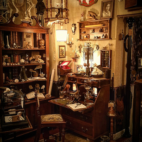 Der Schreibtisch des Doktors.jpg Ein opulenter, historisch eingerichteter Raum mit einem antiken Schreibtisch, Büchern, Lampen und wissenschaftlichen Instrumenten.