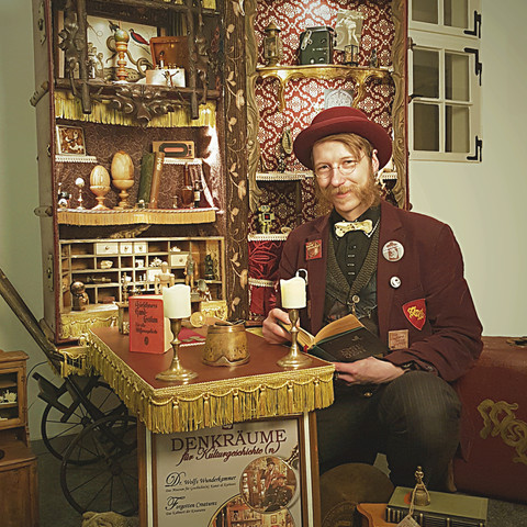 Eine Welt im Reisekoffer - Die Wunderkammer unterwegs.jpg Auf dem Bild ist ein Mann zu sehen, der in einem nostalgisch dekorierten Raum sitzt. Hinter ihm steht ein offener, kunstvoll gestalteter Holzschrank, gefüllt mit kleinen Artefakten und dekorativen Gegenständen. Der Mann trägt ein klassisches Outfits samt Rundbrille und Zylinder, liest in einem Buch und sitzt an einem Tisch, der mit einer Kerzenleuchte geschmückt ist. Der Raum strahlt eine gemütliche, kuriose Atmosphäre einer Wunderkammer aus.