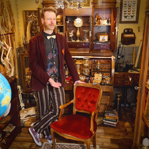 Willkommen in der WK 2024.jpg Ein Mann in einem stilvollen, vintage-inspirierten Raum zeigt ein hohes, rotes Polstersessel. Breite Holzmöbel, ein Globus und skurrile Sammlerstücke zieren den Hintergrund. Sein Outfit mit Fliege und gestreiften Hosen verstärkt den alten Charme des Zimmers.