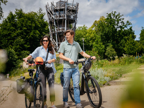 Ein Paar fährt lächelnd auf E-Bikes an einem hohen Aussichtsturm vorbei.