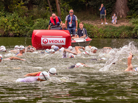 Participants of a triathlon swim in open water towards a buoy, spectators on the shore.