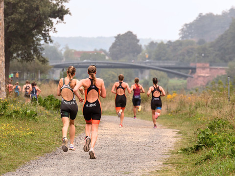 Triathletes run along a wide path, surrounded by trees and countryside, towards the bridge.