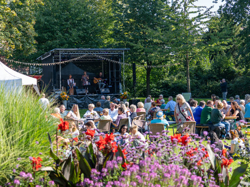 Openluchtconcert op het platteland met muzikanten op het podium en publiek aan tafels in de zon.