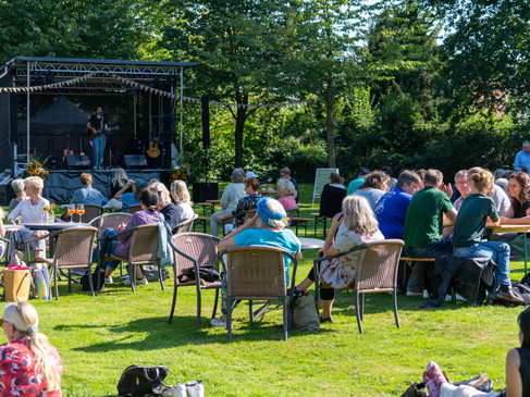 Leute sitzen auf Stühlen im Freien, blicken auf eine Bühne mit Musikern.