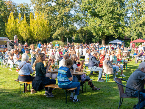 Menschen genießen ein Freiluft-Event im Park bei sonnigem Wetter, sitzend an Holzbänken.