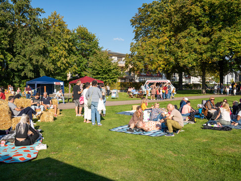 Menschen genießen ein sonniges Picknick im Park, umgeben von Bäumen und mehreren Ständen.