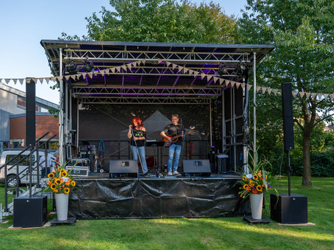 Freiluftbühne mit zwei Musikern unter bunten Wimpeln vor grünem Laub.