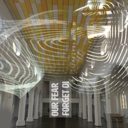 Colours of Peace Eine leuchtende Lichtinstallation projiziert Wellenmuster auf die gelb-weiß gestreifte Decke einer Kirche, die von eleganten weißen Säulen getragen wird. Die Worte "OUR FEAR FORGET" heben sich in klarer Schrift vom dunklen Boden ab.A luminous light installation projects wave patterns onto the yellow and white striped ceiling of a church, which is supported by elegant white pillars. The words "OUR FEAR FORGET" stand out from the dark floor in clear lettering.
